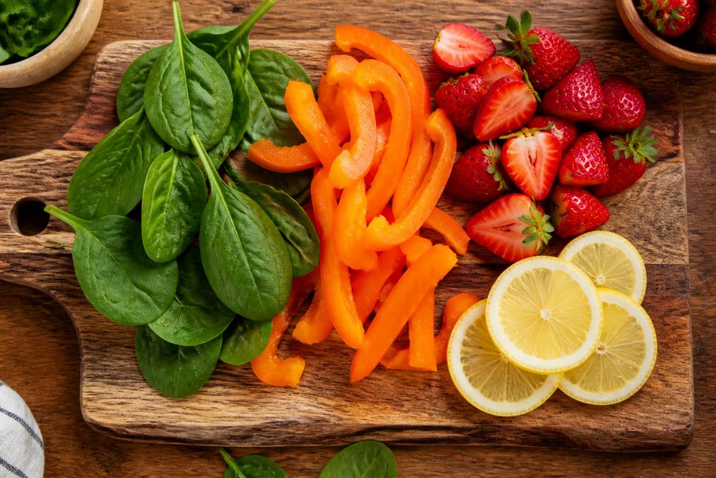 Overhead view of a wooden cutting board with fresh leafy greens, bright orange bell peppers, red strawberries, and citrus lemon slices arranged together, natural kitchen lighting