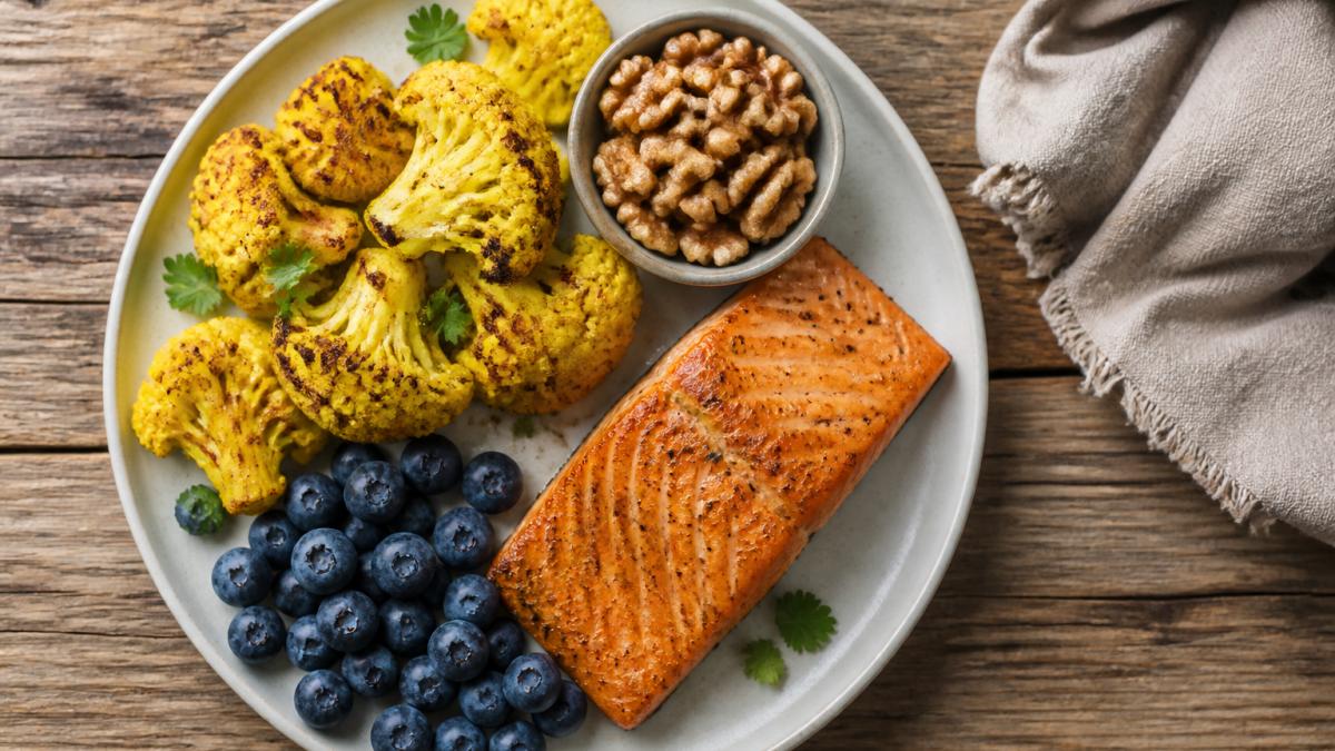 Overhead shot of a colorful anti-inflammatory meal: grilled salmon fillet, roasted turmeric cauliflower, fresh blueberries, and a small bowl of walnuts on a rustic wooden table with natural lighting