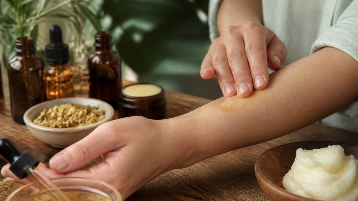 Close-up of hands applying botanical oils and natural moisturizers to arm skin, showing golden oil droplets absorbing into dry patches with warm natural lighting