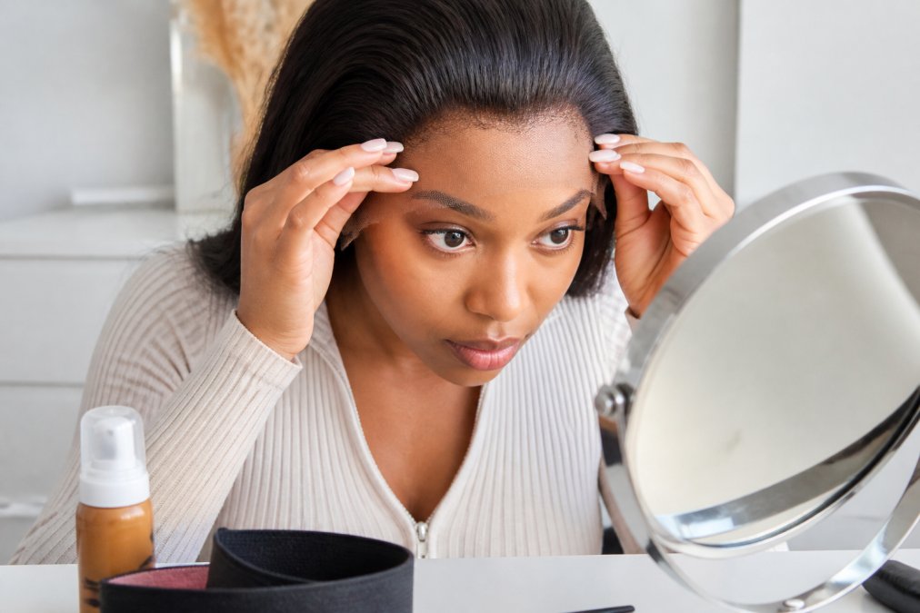 Close-up of a woman's hands gently pressing a lace front wig cap onto her hairline, with a lace tint mousse bottle and wig melt band visible on a clean dressing table nearby