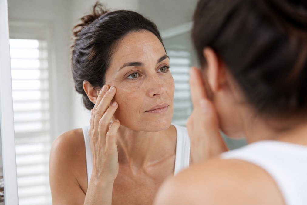 Close-up of a woman in her 40s gently touching her cheek while looking into a bathroom mirror, soft natural daylight streaming through a window, visible signs of dry texture on her skin