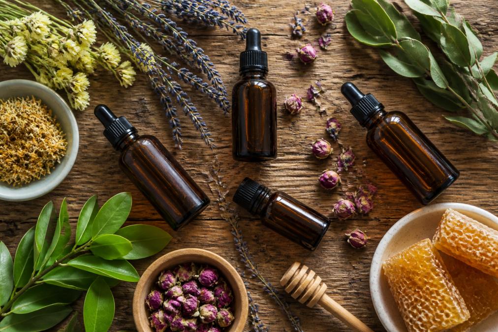 Flat lay of several small amber glass bottles of botanical oils surrounded by dried flowers, honeycomb pieces, and green leaves on a warm wooden surface