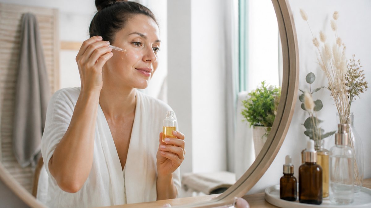 Woman in her 30s applying facial serum at a bright vanity with an ice roller and glass bottles nearby