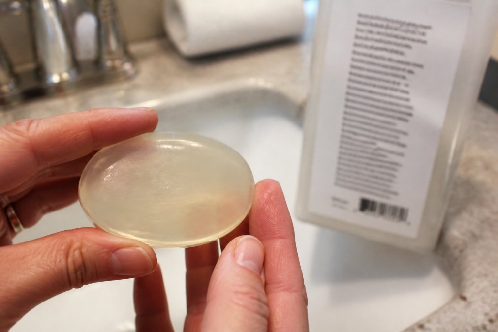 Close-up of hands holding a clear bar of commercial soap, with a list of chemical ingredients visible on the packaging blurred in the background, harsh bathroom lighting