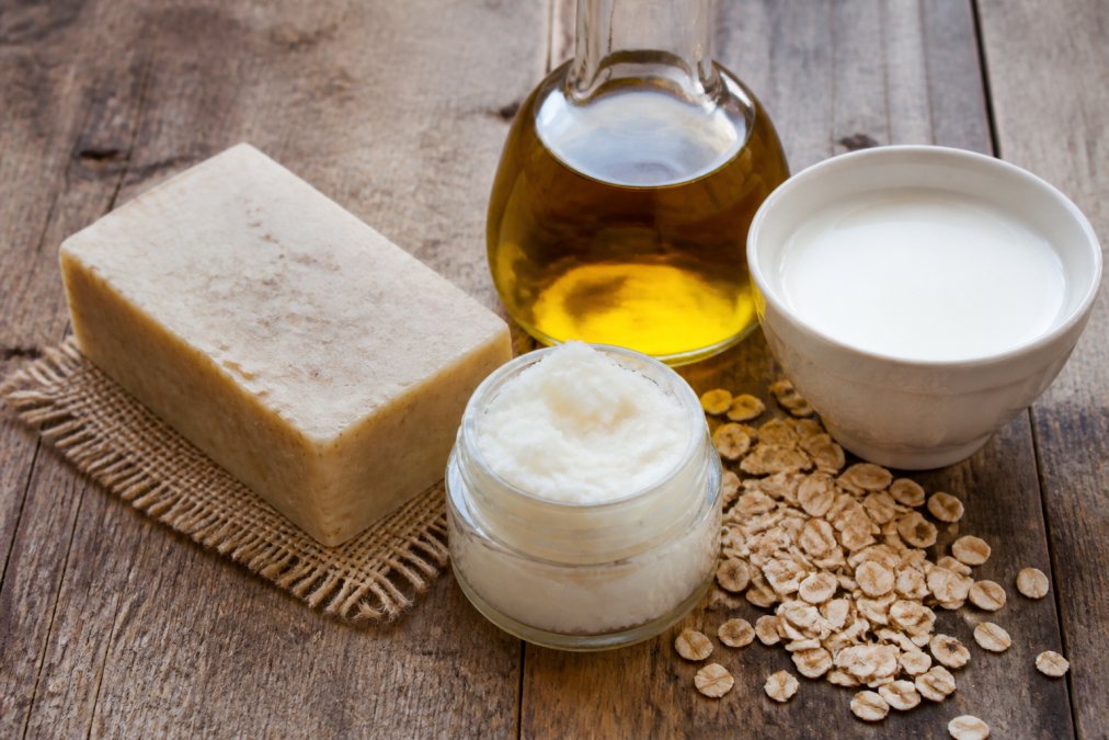 Flat lay of natural soap-making ingredients arranged on a worn wooden surface: a rustic bar of handmade soap, a small open jar of shea butter, olive oil in a clear glass bottle, dried oats scattered loosely, and fresh goat milk in a white ceramic bowl, photographed under warm natural window light