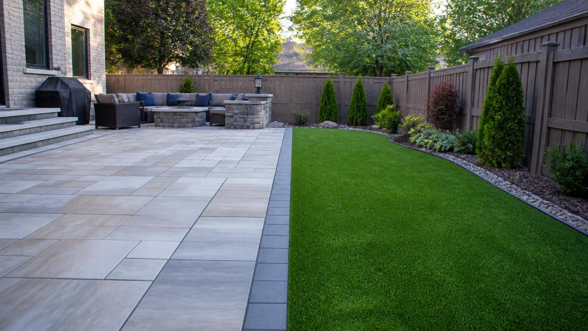 A finished backyard in a Michigan suburb showing a natural stone patio on one side and a strip of artificial turf on the other, with landscaping and a fence in the background