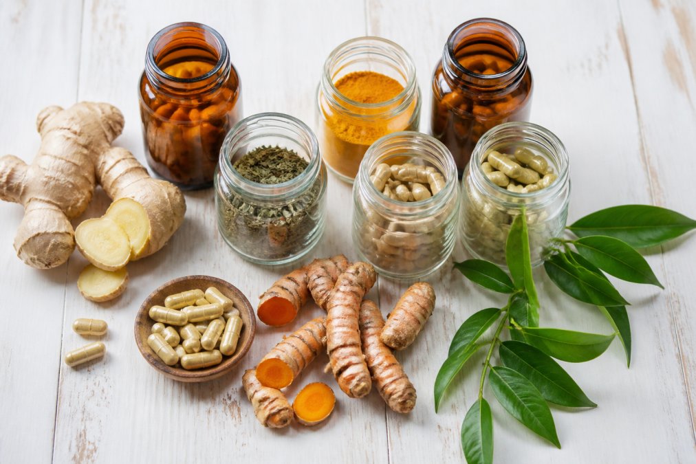 Flat lay of glass jars and amber supplement bottles alongside fresh turmeric root, ginger, and green tea leaves on a white wooden surface in natural daylight