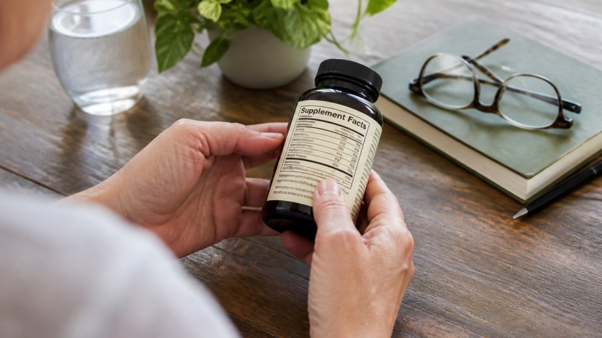 Close-up of hands holding a supplement bottle while reading the label in natural light, with glasses and a wellness journal nearby on a wooden table