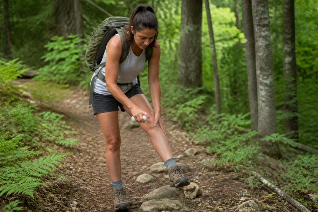 Hiker applying natural spray repellent to legs and exposed skin before heading into wooded trail, demonstrating proper coverage technique