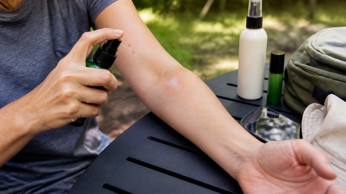 Close-up of a person applying a small patch of tick spray to the inside of their forearm, with a timer or phone nearby on an outdoor table, natural daylight