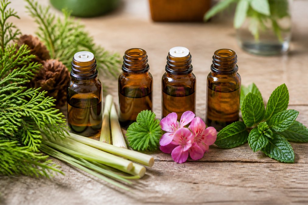 Close-up of amber glass bottles containing lemongrass, cedarwood, geranium, and peppermint essential oils arranged on a natural wooden surface with fresh botanical sprigs