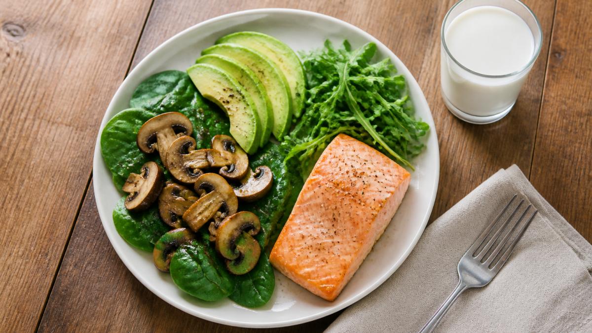 Overhead shot of a colorful plate with leafy greens, mushrooms, avocado, salmon, and a glass of milk on a wooden table, natural daylight
