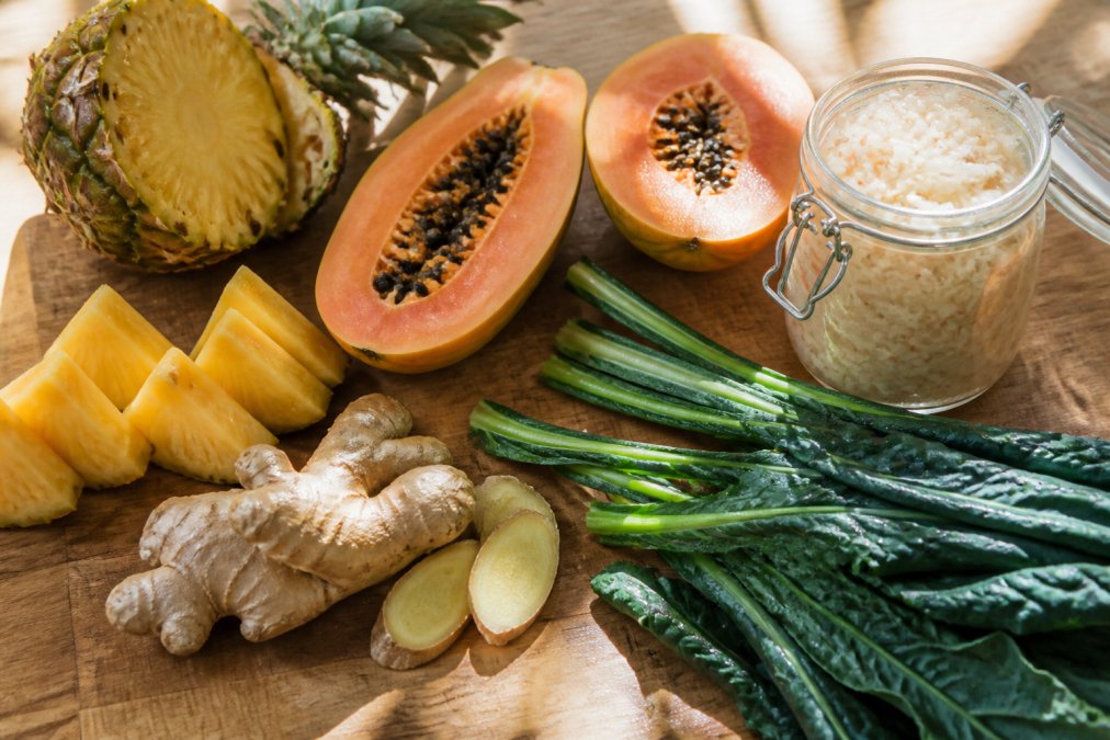 Overhead shot of fresh foods on a wooden cutting board including sliced pineapple, papaya halves, fresh ginger root, fermented sauerkraut in a glass jar, and dark leafy greens with natural sunlight streaming across the surface