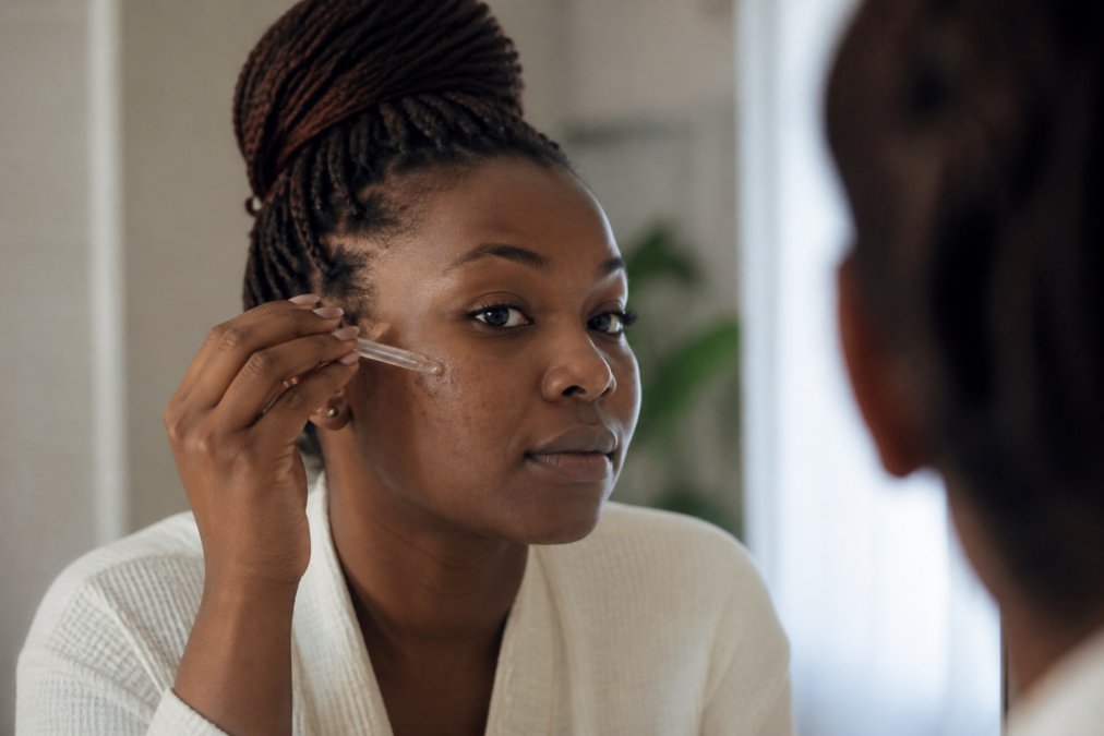 Close-up of a Black woman with sensitive skin gently applying a serum to her cheek in a bright bathroom, soft natural light, calm and focused expression