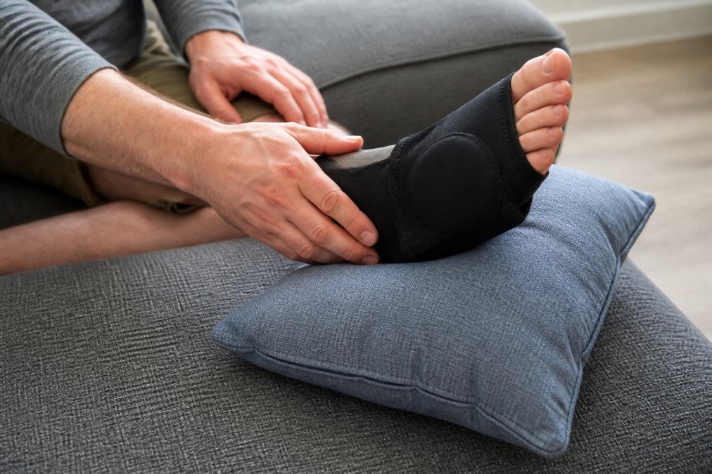 Close-up of a person applying a cold compression sleeve to a swollen big toe and foot joint while resting on a couch, foot propped on a firm pillow, soft indoor lighting