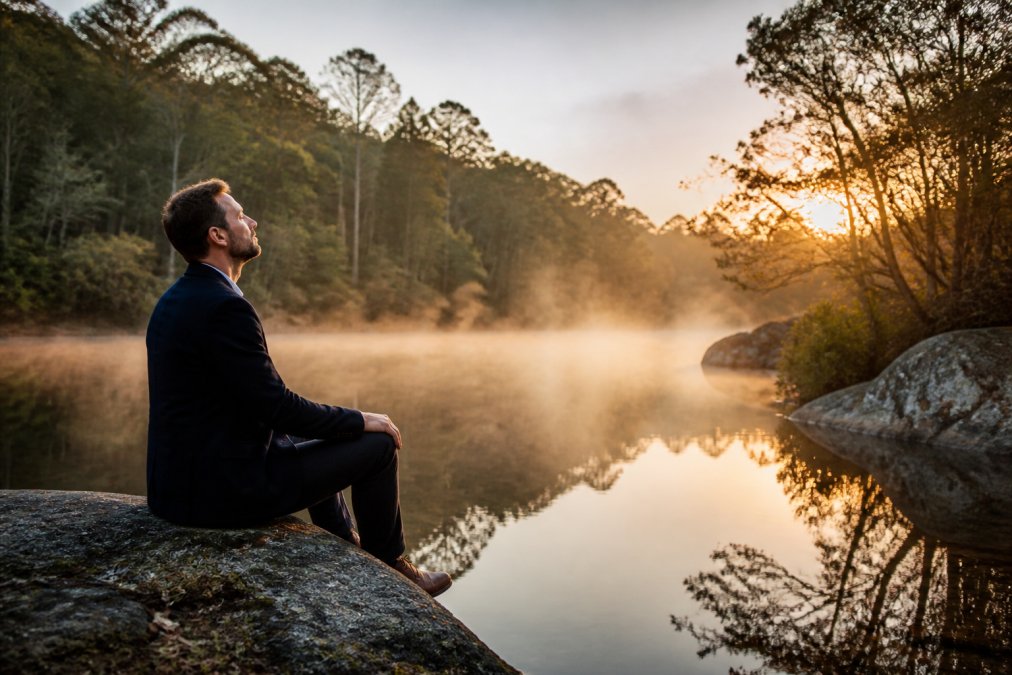 Corporate leader sitting alone by a natural water feature at dawn, shoulders relaxed, breathing deeply with peaceful expression, morning mist rising from the water