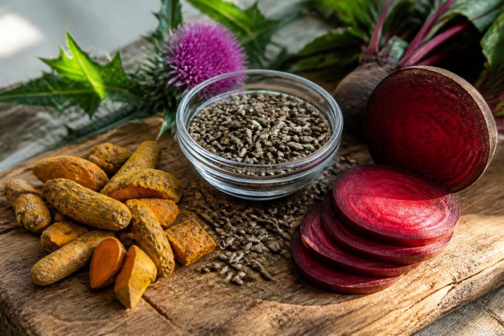 Close-up of natural ingredients including dried turmeric root chunks, milk thistle seeds in a small glass bowl, and sliced fresh beet root on a weathered wooden cutting board with soft morning light streaming across the surface