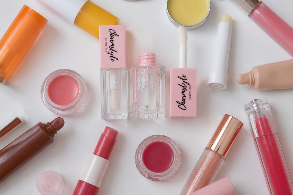 Overhead flat lay of various lip balm and lip gloss products arranged on a clean white surface, showing different textures, colors, packaging styles, and formulations