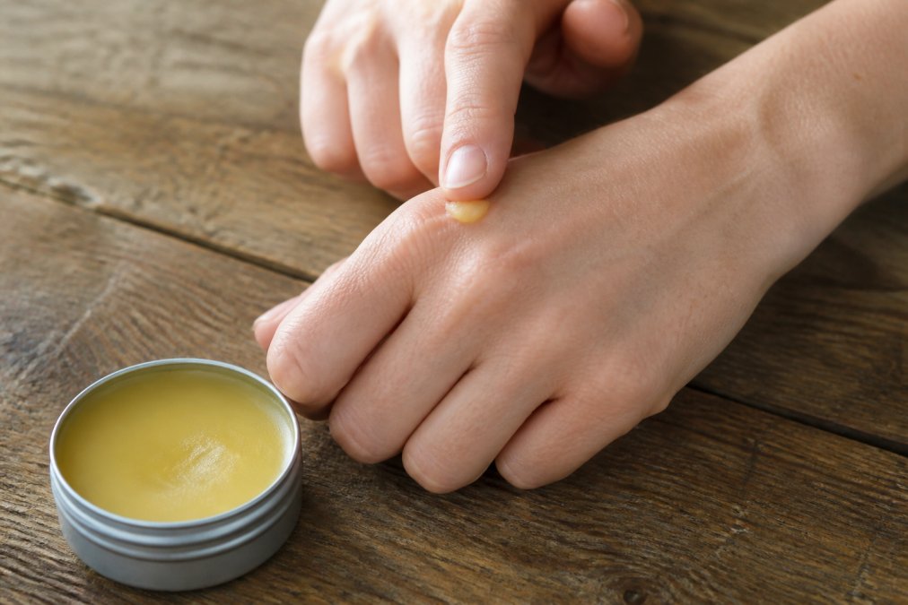 Close-up of hands applying a small amount of balm from an open tin container onto dry skin, natural daylight, rustic wooden table background