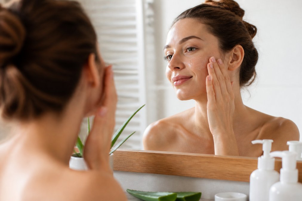 Close-up of a woman's hands gently applying a clear aloe vera gel to her cheek in front of a bright bathroom mirror, soft morning light