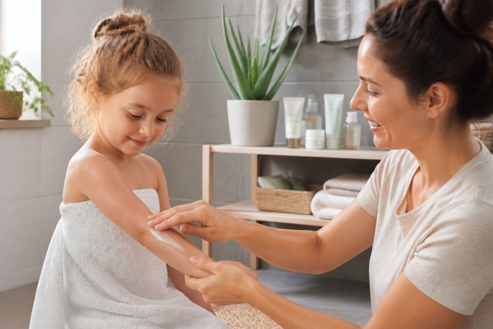 Barn der får påført mild fugtighedscreme på armen af en voksen i et lyst badeværelse med naturlige plejeprodukter på hylden