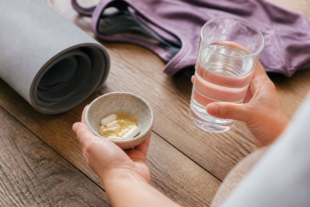 Close-up of hands holding a glass of water and a small bowl with vitamin capsules on a wooden table, yoga mat and athletic clothing softly blurred in the background, bright natural daylight