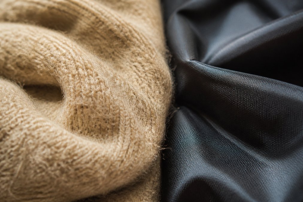 Nahaufnahme einer feinen Wollstruktur mit sichtbaren Fasern in warmem Licht, daneben ein glänzendes synthetisches Textil, das den Kontrast in der Oberflächenstruktur deutlich zeigt