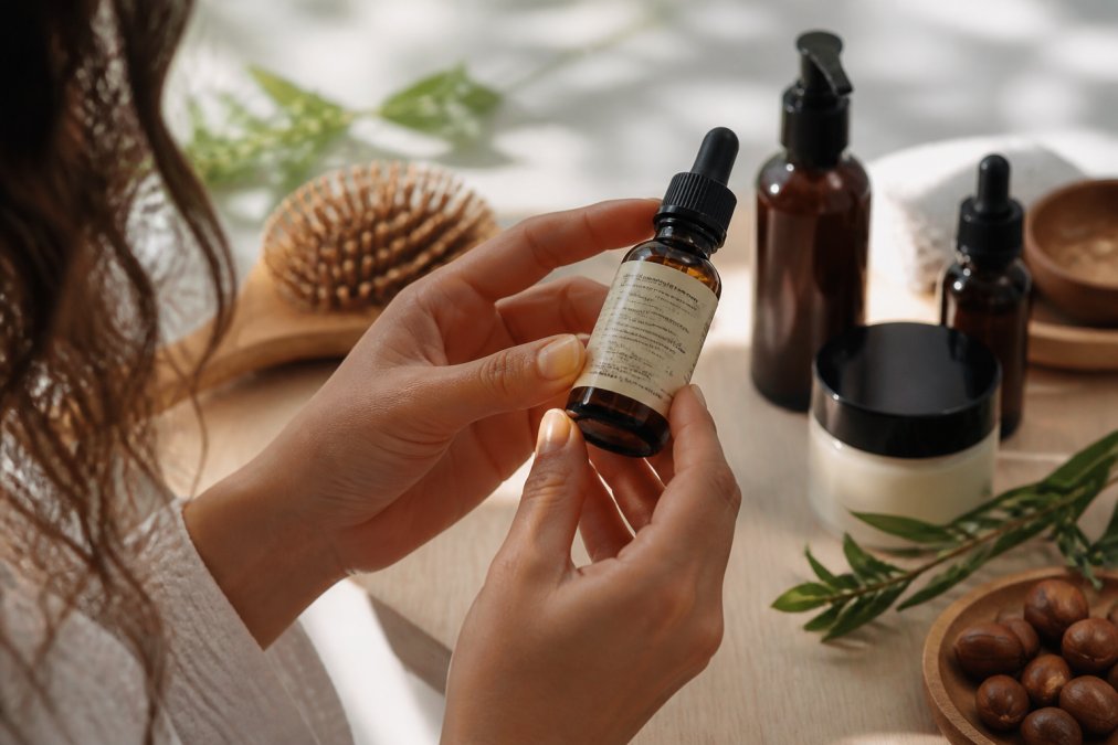 Close-up van een hand die een glazen flesje met natuurlijke haarolie vasthoudt en aandachtig de ingrediëntenlijst op het etiket bestudeert, met zachte daglichtinval en andere natuurlijke verzorgingsproducten op de achtergrond