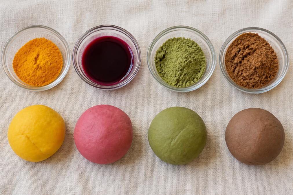 Flat-lay of small glass bowls containing natural colorants, turmeric powder, beetroot juice, spinach powder and cocoa, arranged next to balls of freshly made clay in matching colors on a light linen surface, photographed from directly above