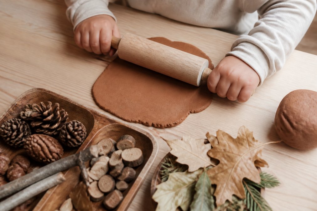 Close-up van kinderhanden die natuurlijke speelklei uitrollen met een houten roller, met een houten speeltray en losse natuurmaterialen zoals dennenappels en bladeren ernaast op een lichte houten tafel