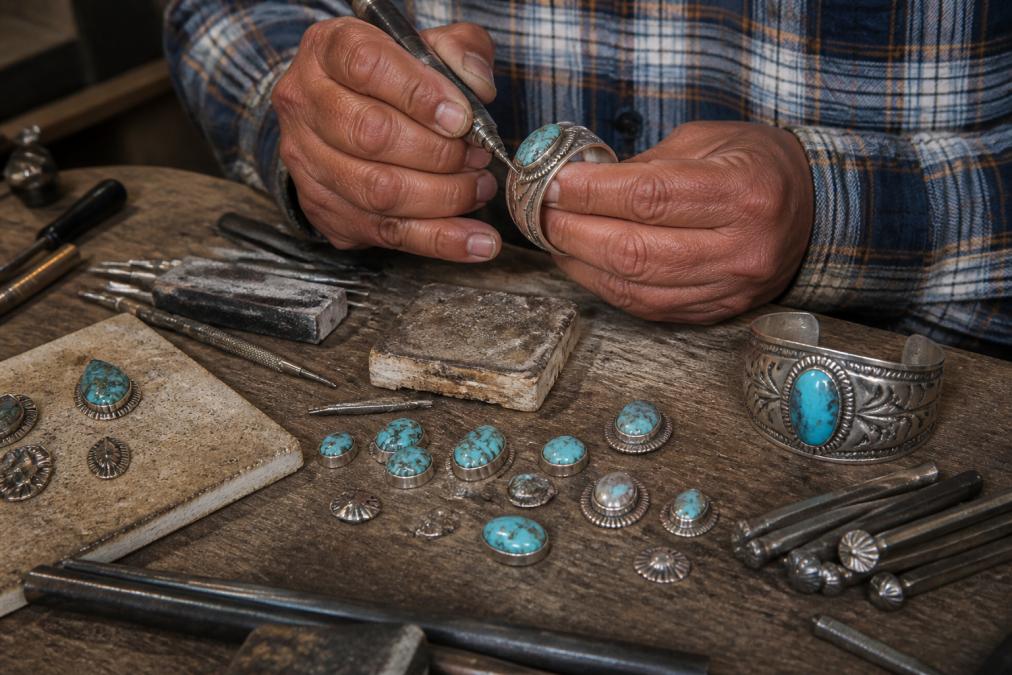 Close-up of artisan hands crafting sterling silver Navajo jewelry with turquoise stones on a workbench, showing traditional silversmith tools and materials