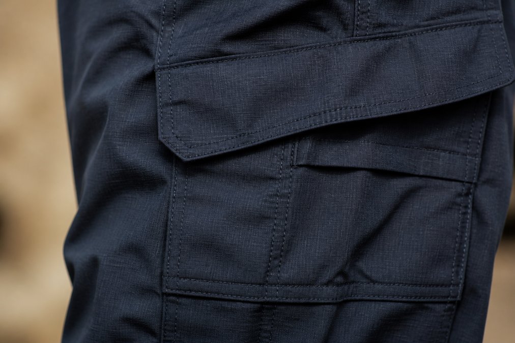 Close-up of ripstop fabric weave on navy tactical pants showing reinforced stitching, utility pockets, and durability details under studio lighting