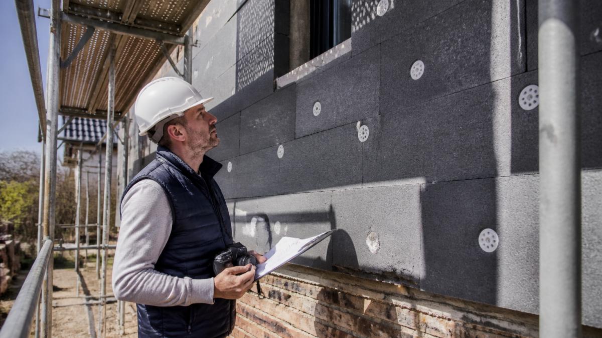 Vastgoedbeheerder die tijdens inspectie een gevelisolatie controleert met checklist en fotocamera in de hand op een renovatieproject in volle daglicht