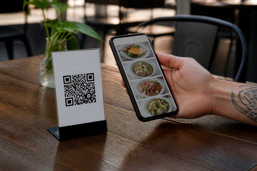 Código QR impreso en una tarjeta de mesa de restaurante junto a un smartphone mostrando el menú digital con platos visibles, sobre madera oscura