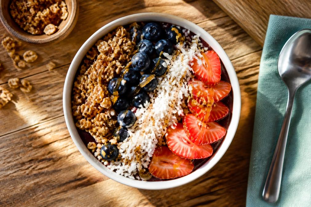 Overhead view of a vibrant açaí bowl topped with granola clusters, fresh blueberries, sliced strawberries, shredded coconut, and honey drizzle on a rustic wooden table with soft morning light streaming across