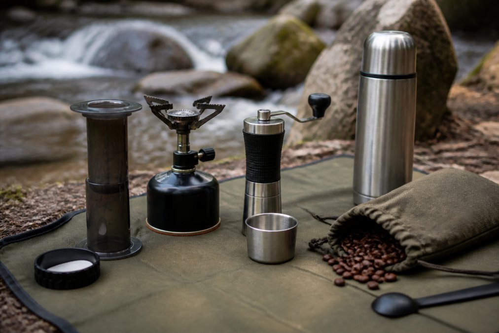 Outdoor coffee brewing equipment neatly arranged on a camping mat in natural daylight, including AeroPress, portable burner, hand grinder, thermos, and coffee beans in a canvas pouch