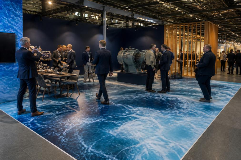 Moderne messestand med farverigt trykt gulv viser virksomhedslogo og grafik, besøgende går over det og fotograferer det, professionel messebelysning