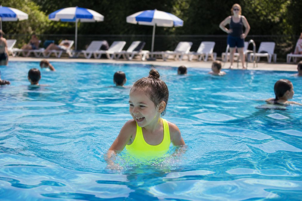 Young child wearing a bright neon swimsuit playing in a busy public pool with other swimmers in the background, parent visible supervising from poolside