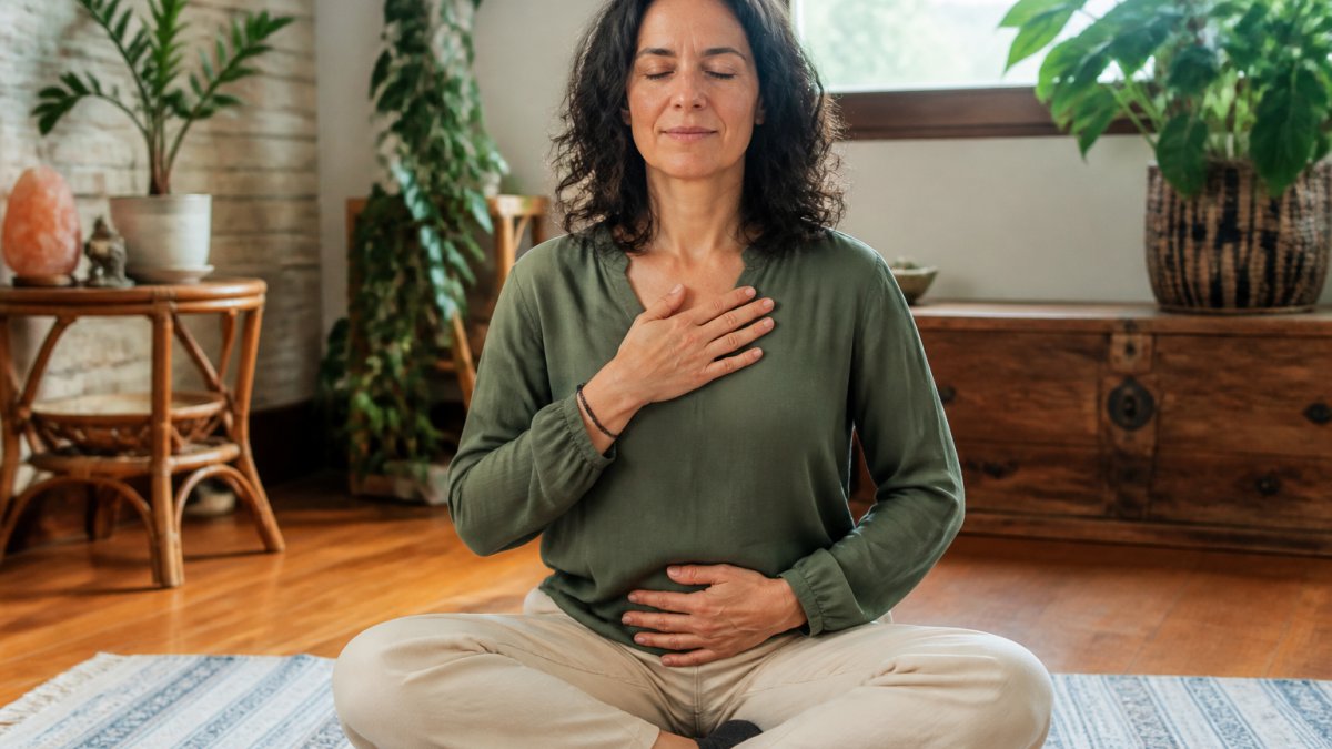 Woman in her 40s sitting cross-legged in a bright, naturally lit room with soft window light, hand on her heart, eyes gently closed, engaged in conscious body awareness practice