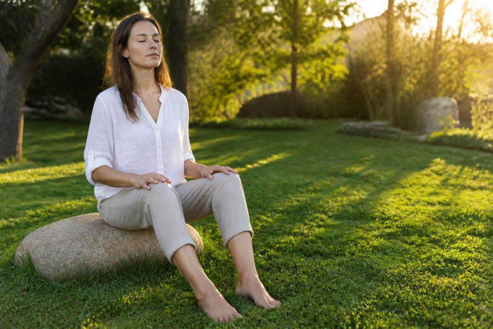 Person sitting outdoors in a calm, natural setting during golden hour, bare feet pressed into grass, hands resting on thighs, with eyes closed and peaceful expression showing embodied presence and grounding