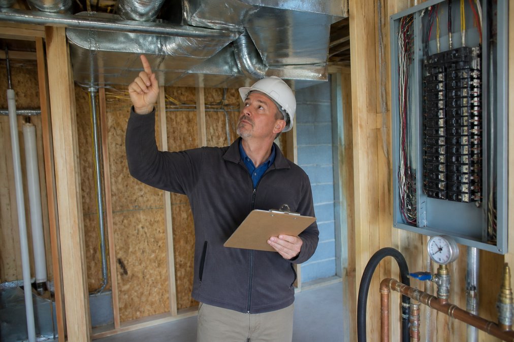Home inspector checking HVAC ductwork connections, electrical panel breakers, and plumbing water pressure in unfinished interior space with equipment and clipboard
