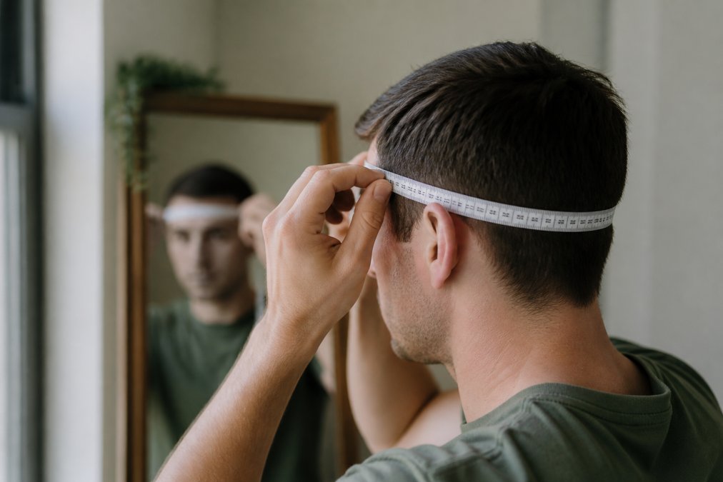 Close-up of a person using a flexible measuring tape around their head, positioned just above the ears, with a mirror in the background and soft indoor lighting