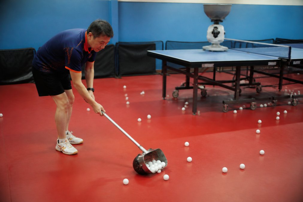 A table tennis coach using a handheld scoop to quickly collect dozens of ping pong balls from the floor of a training facility, with a robot and table visible in the background