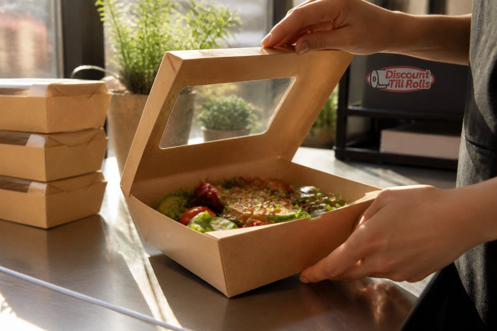 Close-up of hands opening a compostable kraft clamshell food box with windowed lid, showing fresh salad inside, on a café counter with morning light streaming across the workspace