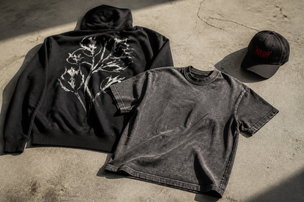Eine Flatlay-Aufnahme der neuesten HTT-Streetwear-Artikel auf einem Betonboden: ein schwarzer Hoodie mit auffälligem Rückendruck, ein graues Snow-Wash-T-Shirt und eine schwarze Cap mit roter Stickerei, beleuchtet von hartem Seitenlicht, das die Textur der Stoffe betont