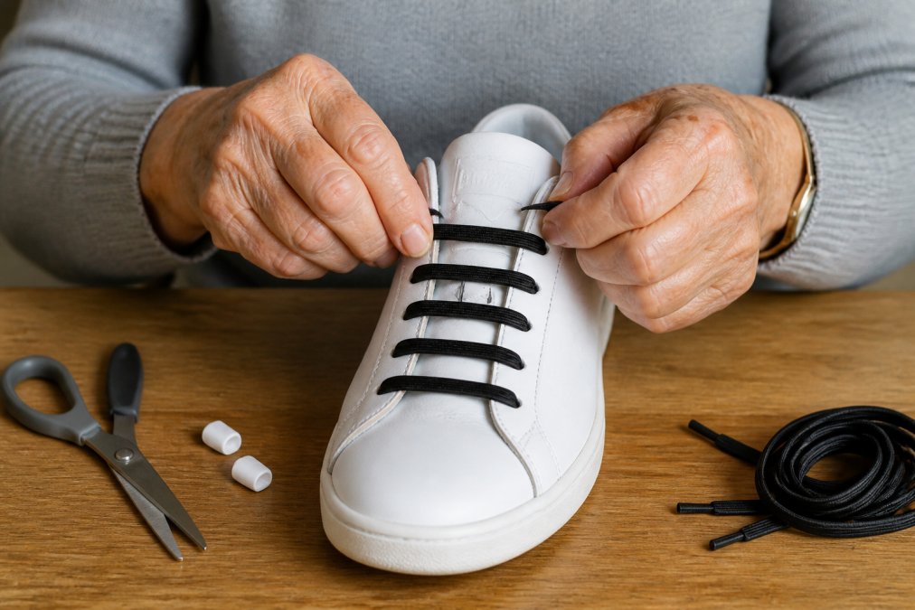 Close-up of hands with visible age spots carefully threading elastic no-tie laces through shoe eyelets, demonstrating the push-and-pull method with clear finger positioning and relaxed hand posture