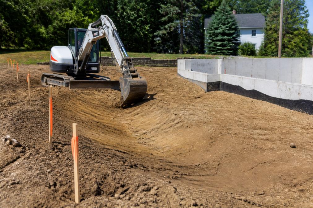 A compact excavator shaping a drainage swale on a residential lot, with surveyor's stakes in the foreground and a house foundation in the background under bright daylight