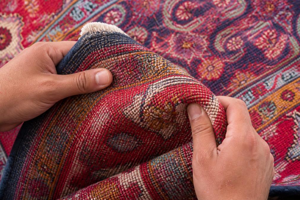 Close-up of hands examining the back of a handwoven kilim rug, showing intricate knot patterns and natural fiber texture under warm studio lighting