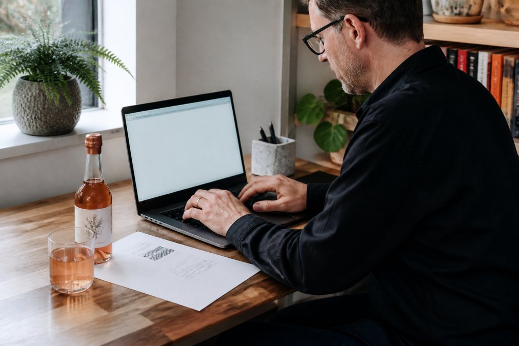 Person writing an email about a non alcoholic drink return policy while sitting at a wooden desk with a delivery note.
