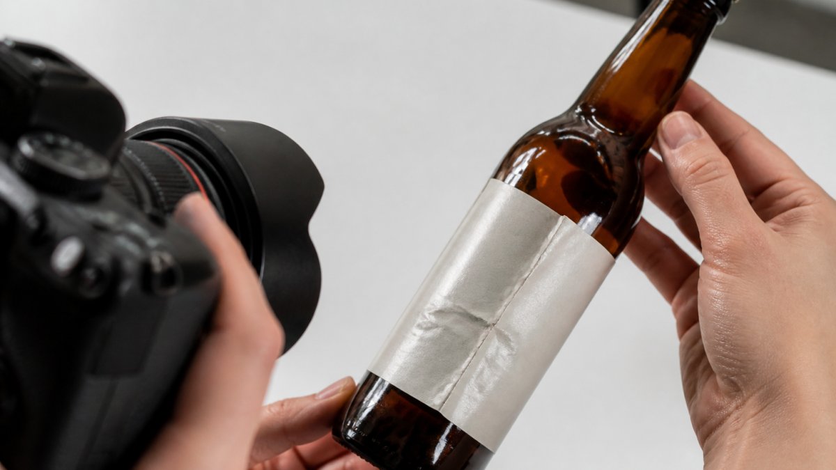 Close-up of a damaged alcohol-free beer bottle with visible dent and label damage being photographed on a white surface with clear natural lighting, showing the damage clearly in focus